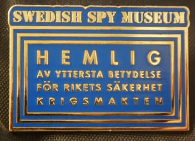 Rektangulär emaljpin i blått med guldfärgad text och tre inramande linjer, formad som en hemligstämpel: SWEDISH SPY MUSEUM i en list överst och HEMLIG · AV YTTERSTA BETYDELSE FÖR RIKETS SÄKERHET · KRIGSMAKTEN inuti ramen.