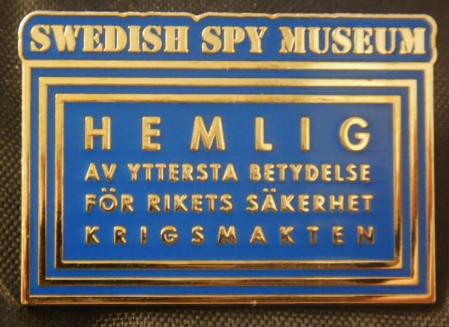 Rektangulär emaljpin i blått med guldfärgad text och tre inramande linjer, formad som en hemligstämpel: SWEDISH SPY MUSEUM i en list överst och HEMLIG · AV YTTERSTA BETYDELSE FÖR RIKETS SÄKERHET · KRIGSMAKTEN inuti ramen.