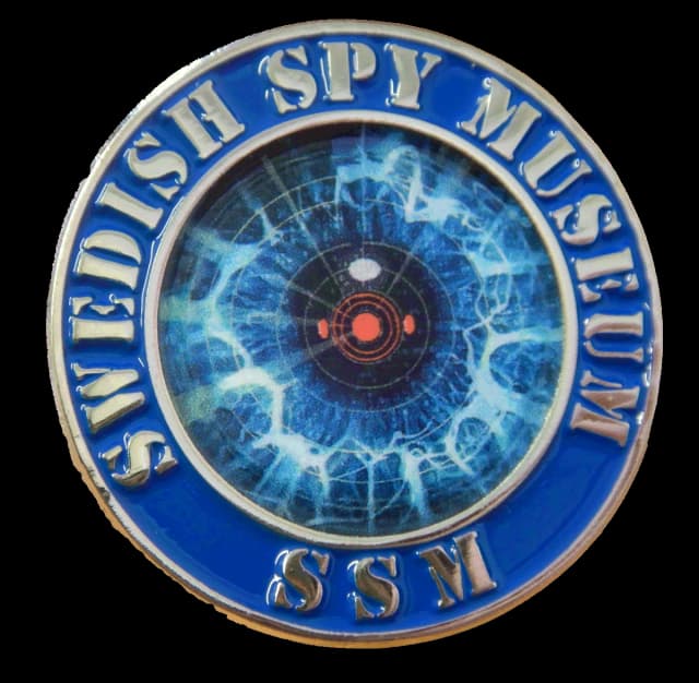 Rund emaljpin i blått med upphöjd silverfärgad text SWEDISH SPY MUSEUM och SSM runt ett radarliknande öga i blått med röd mittpunkt.