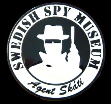 Rund emaljpin i svart och vitt: en silhuett av en spion i bredbrättad hatt och mörka glasögon som håller upp en pistol i handen intill ansiktet, omgiven av texten SWEDISH SPY MUSEUM och namnteckningen Agent Skáti.