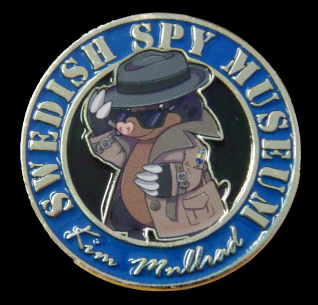 Rund blå emaljpin med den tecknade mullvaden Kim Mullvad i grå hatt, solglasögon och ljus trenchcoat, omgiven av den silverfärgade texten SWEDISH SPY MUSEUM och namnteckningen Kim Mullvad.